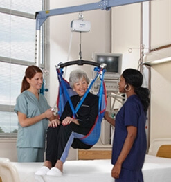 Woman in hospital room in a cieiling lift being helped by 2 nurses