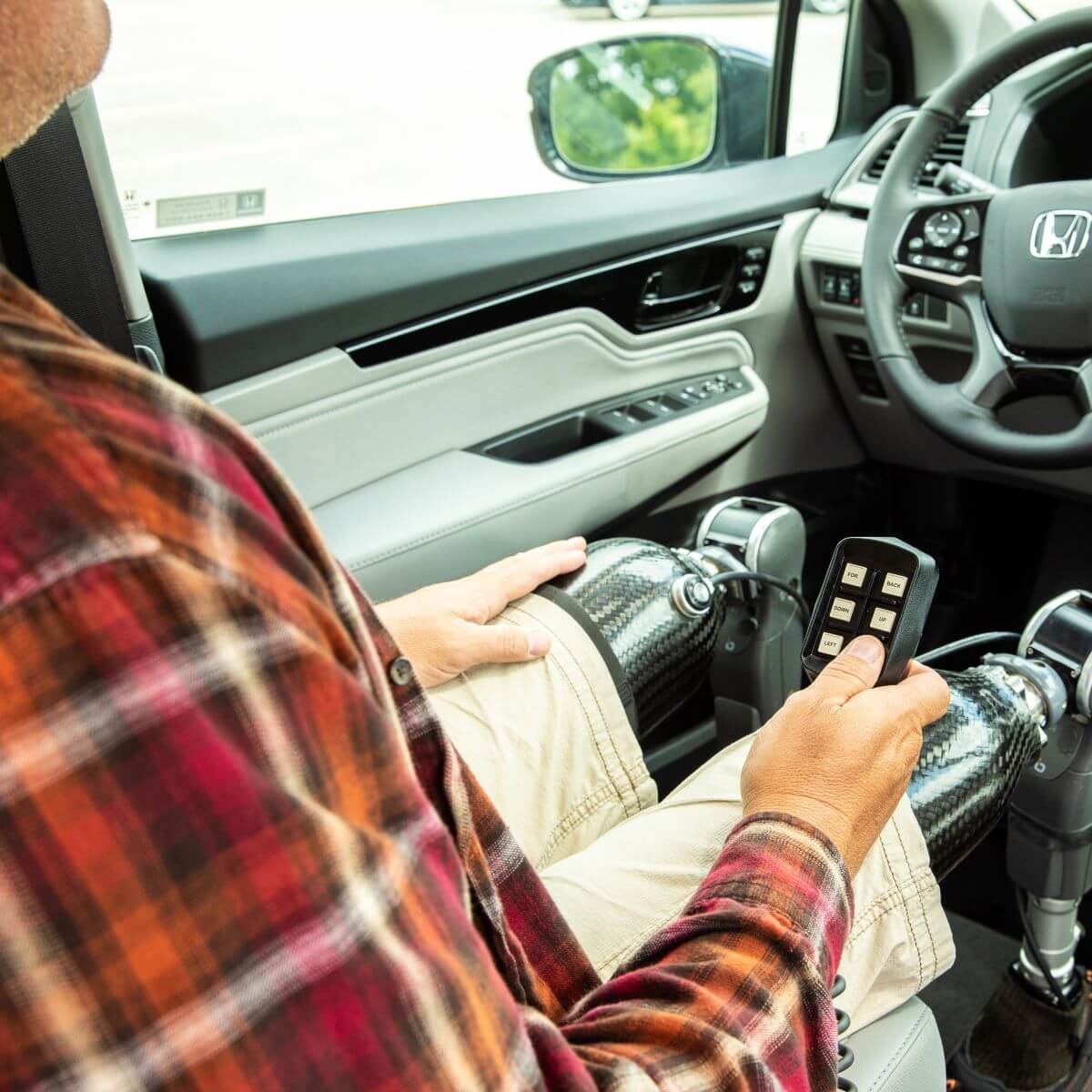 A person with prosthetic legs sits in the drivers seat of a car, holding a remote control device, with the car door open.