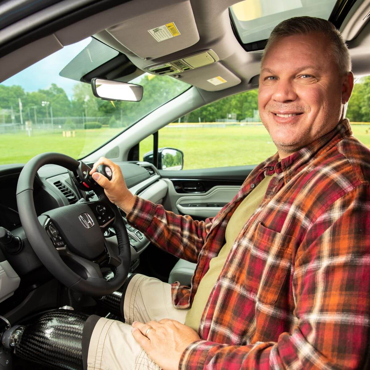 A man with a prosthetic leg sits in the driver’s seat of a car, smiling, with his hand on the steering wheel. A park is visible through the window.
