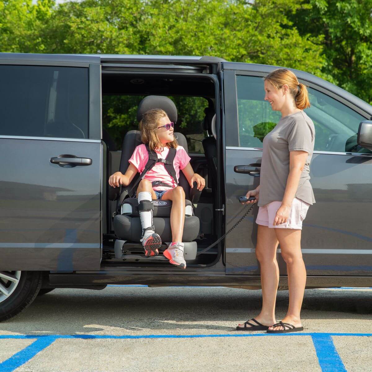 A woman uses a remote control to operate a lift seat, helping a young girl with a leg brace exit a minivan in a parking lot.