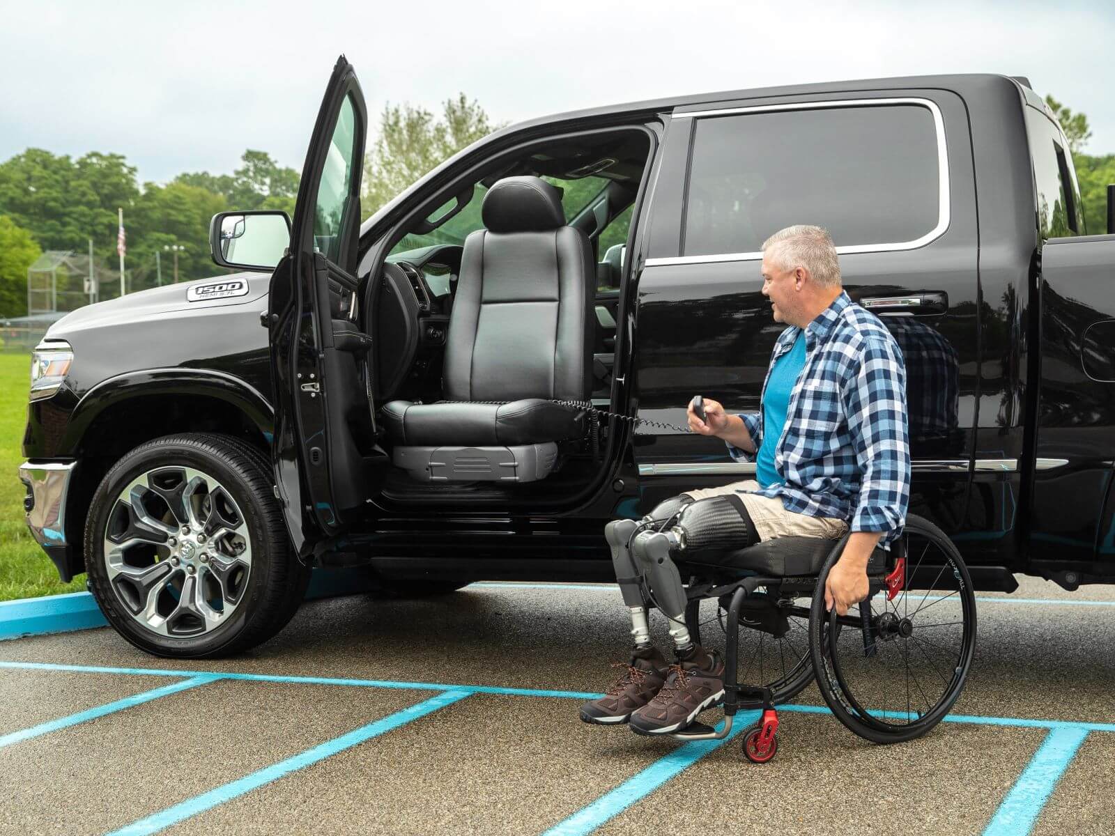 A man in a wheelchair with prosthetic legs uses a remote to operate a modified truck featuring transfer seats and a rotating driver's seat in an accessible parking space.