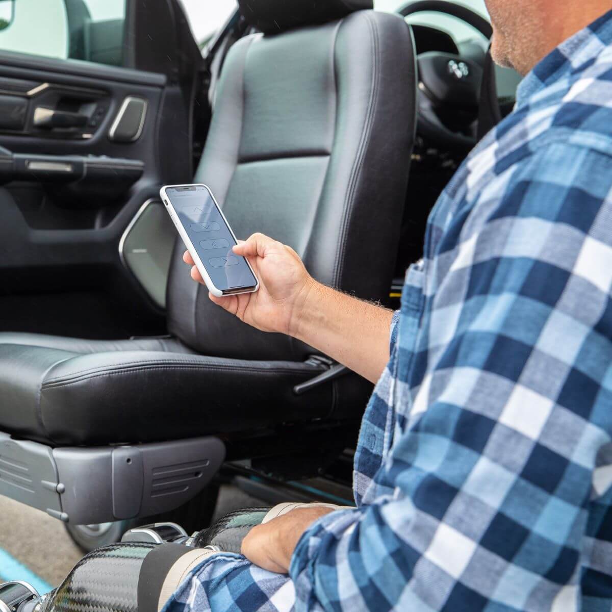 A person in a plaid shirt uses a smartphone while sitting next to an open car door with a swivel seat.