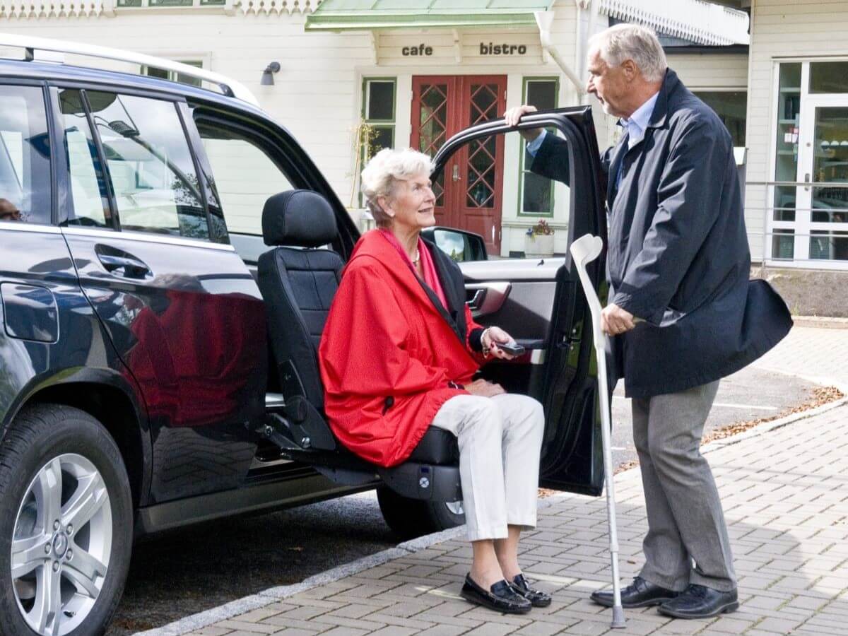An older woman sits on a swivel car seat while an older man with a cane stands beside her in a parking lot outside a cafe and bistro.