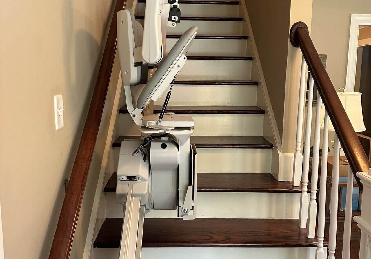 1722633397489 A stair lift installed on a wooden staircase with beige walls and a wooden bannister.