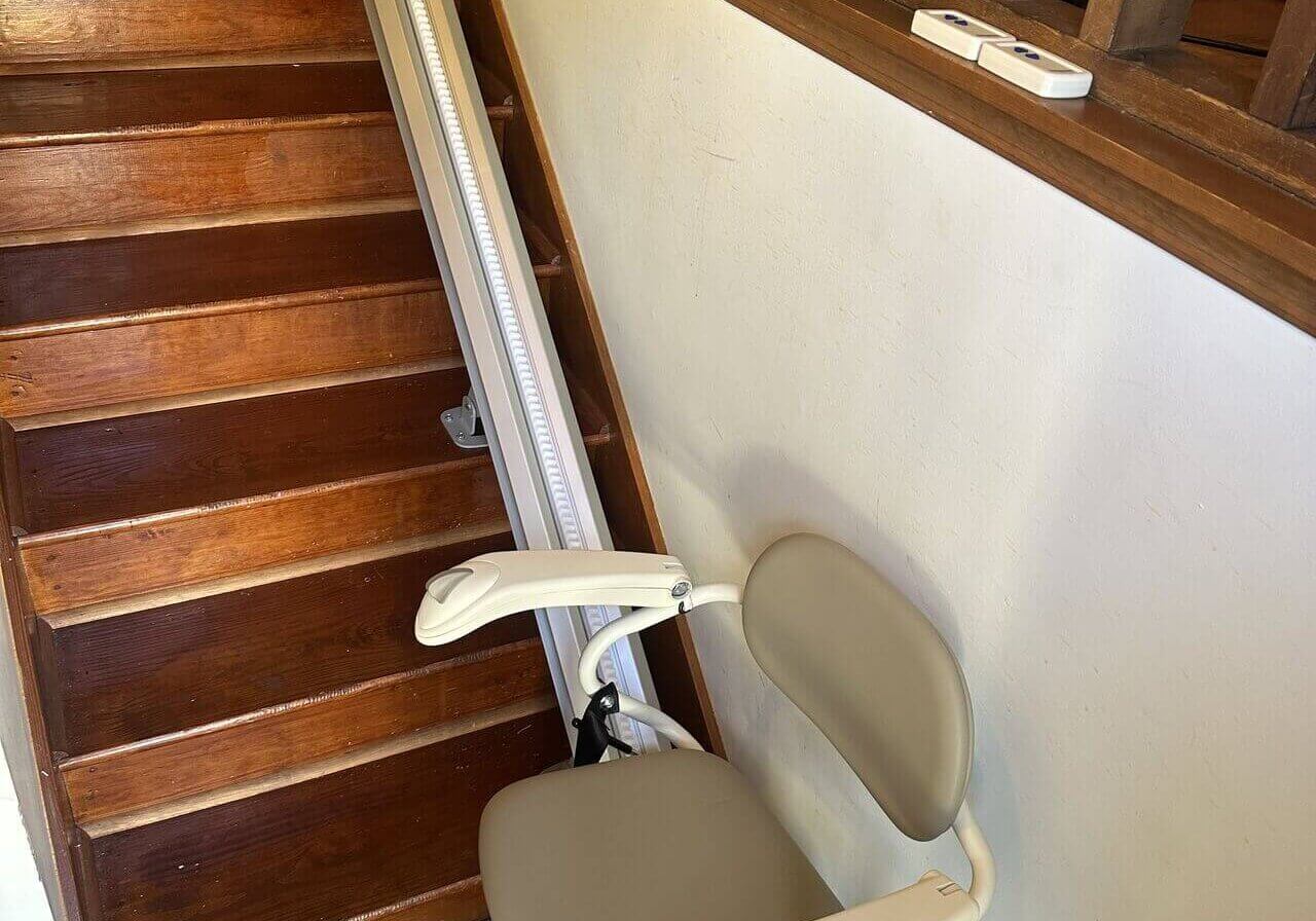 1733507534512 Stair lift chair installed on a wooden staircase with a beige seat and safety belt.