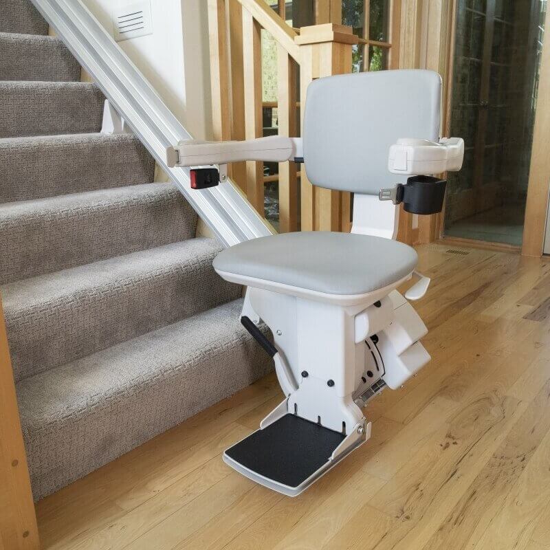 bruno-elite-straight-stair-lift-2 A stairlift is installed alongside a residential staircase, featuring a gray seat and armrests.