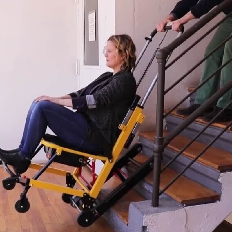 mobilestairlift A person uses a yellow, mobile stair climbing wheelchair to descend stairs, assisted by another person controlling the chair from behind.