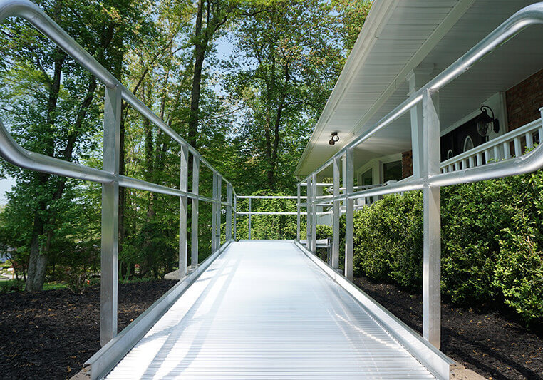 A metal accessibility ramp leads to the entrance of a house surrounded by greenery, seamlessly complementing the stairlifts inside.